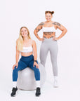 Two women in sports bras and leggings striking a pose on a ball. Female activewear.