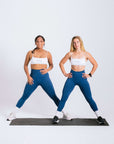 Two women in matching blue leggings and white sports bras workout gear standing on a mat, getting ready to exercise.