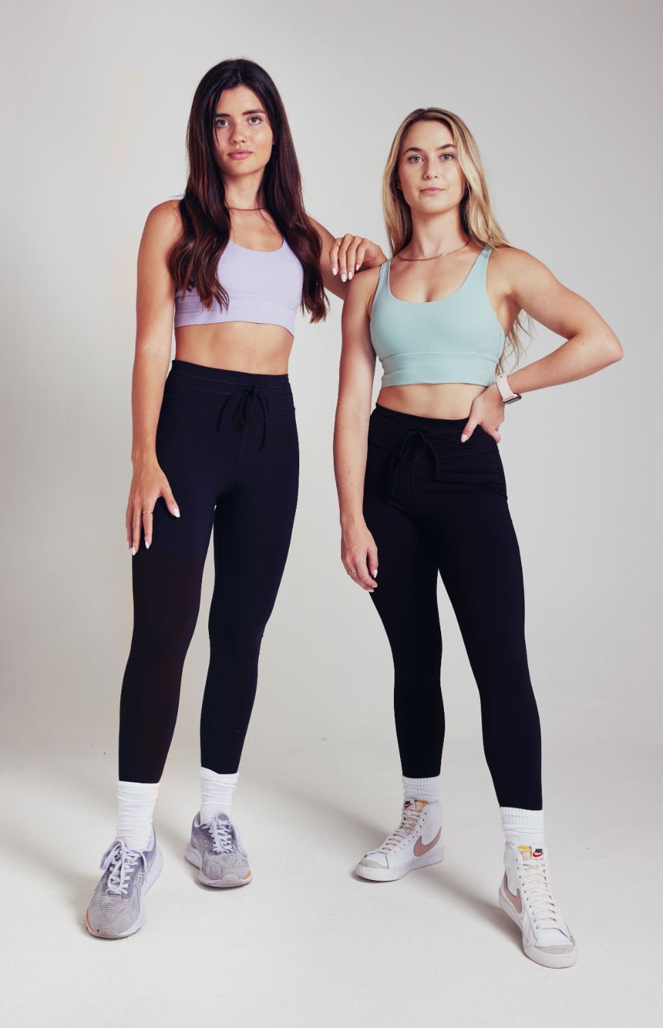 Two fit women standing in black drawstring leggings and pastel coloured sports bras.