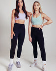 Two fit women standing in black drawstring leggings and pastel coloured sports bras.